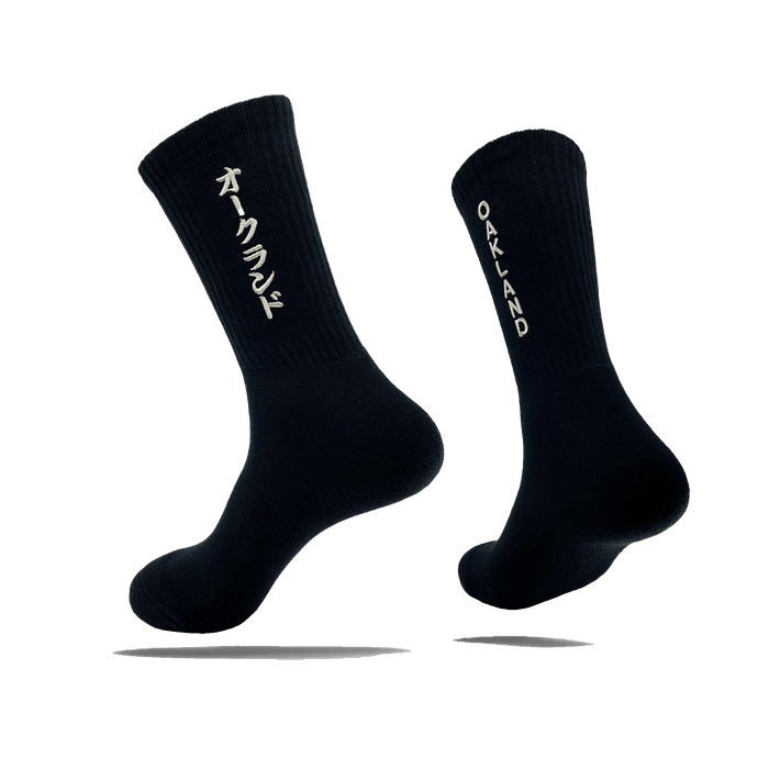 Side view of a pair of black crew socks with OAKLAND wordmark on the side of one sock and Kanji Japanese writing on the other.