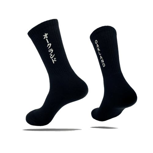 Side view of a pair of black crew socks with OAKLAND wordmark on the side of one sock and Kanji Japanese writing on the other.