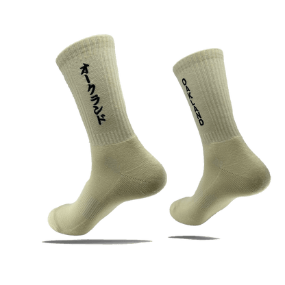 Image of the "Kanji Sock" by Oaklandish, a pair of off-white, ribbed ankle socks. The left sock displays Japanese Katakana characters in black, while the right sock features the word "OAKLAND" in black capital letters. Both cotton socks are shown against a plain white background, slightly elevated to showcase their design. Available in adult sizes.