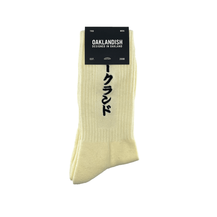 Product Description: The Kanji Sock by Oaklandish is a pair of cream-colored cotton crew socks featuring vertical Japanese characters. A black label at the top reads "OAKLANDISH" with "DESIGNED IN OAKLAND" below it. The label also includes "TRD MRK," "EST.," "2000," and a tree logo. Available in adult sizes.