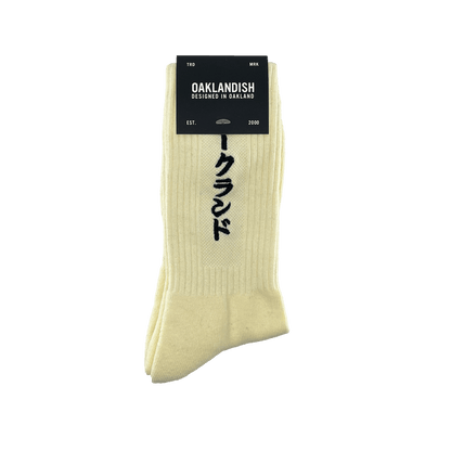 Product Description: The Kanji Sock by Oaklandish is a pair of cream-colored cotton crew socks featuring vertical Japanese characters. A black label at the top reads "OAKLANDISH" with "DESIGNED IN OAKLAND" below it. The label also includes "TRD MRK," "EST.," "2000," and a tree logo. Available in adult sizes.