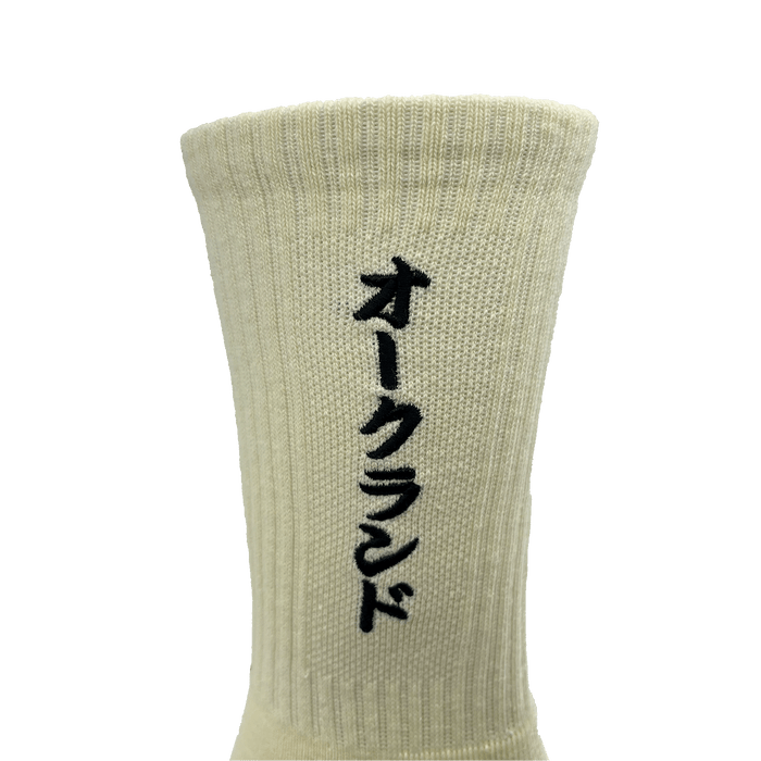 Close-up image of the Oaklandish Kanji Sock in beige cotton, featuring vertical black Japanese katakana characters embroidered on it. The crew sock appears ribbed and has a distinct vertical stretch. Available in adult sizes, the plain white background highlights the texture and color of this stylish sock.