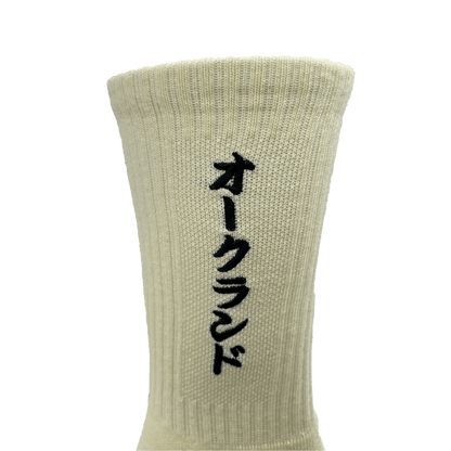 Close-up image of the Oaklandish Kanji Sock in beige cotton, featuring vertical black Japanese katakana characters embroidered on it. The crew sock appears ribbed and has a distinct vertical stretch. Available in adult sizes, the plain white background highlights the texture and color of this stylish sock.