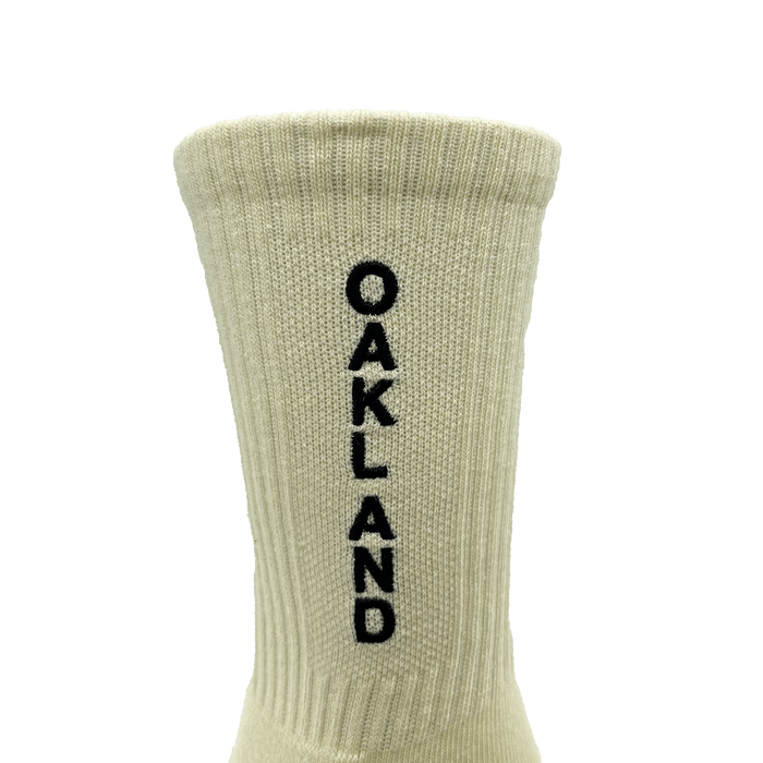 The Kanji Sock by Oaklandish is a beige ribbed cotton crew sock with "OAKLAND" stitched in black vertically down the back. Available in adult sizes, it features a cuff at the top and is set against a plain white background.
