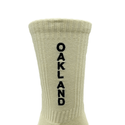 The Kanji Sock by Oaklandish is a beige ribbed cotton crew sock with "OAKLAND" stitched in black vertically down the back. Available in adult sizes, it features a cuff at the top and is set against a plain white background.