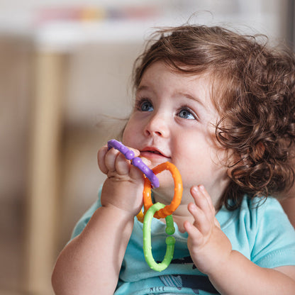 A toddler with curly brown hair and blue eyes wears a blue shirt and holds Nuby Linkables Teething Toys - Primary Colors (18 pack), bringing them to their mouth while looking up.