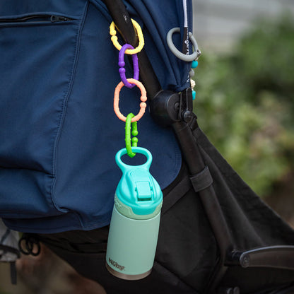 A mint green water bottle hangs from a stroller using Nuby Linkables Teething Toys - Primary Colors - 18 pack attached to the handle. The dark blue and black stroller stands out against a blurred green background.