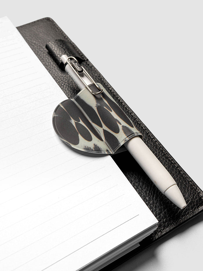 Magnetic Bookmark | Pen Holder | Vinyl in Dissolve holding a white pen, clipped to the spine of an open black leather planner