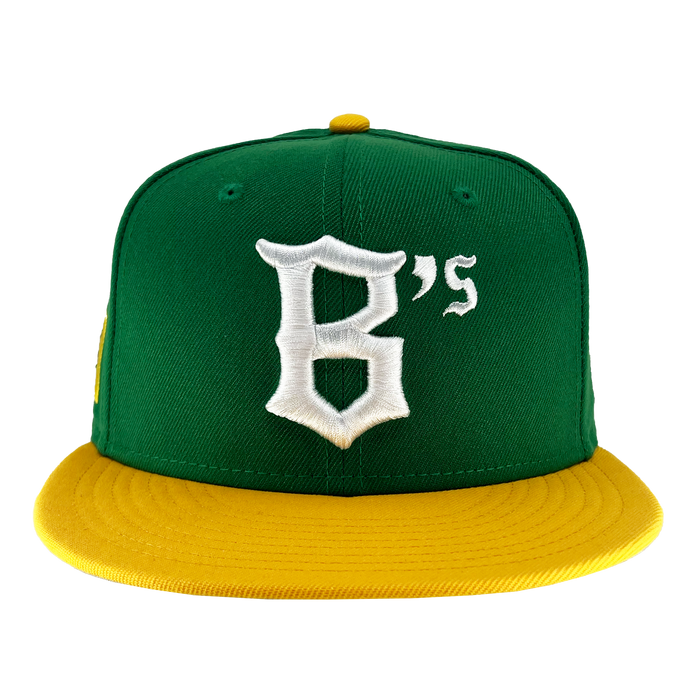 The New Era Oakland Ballers 59Fifty Cap features a green body with a flat yellow brim and a top button that matches in yellow. The front of the cap showcases a white, stylized letter "B" followed by a smaller white "'s," symbolizing the Oakland Ballers. The image background is white.