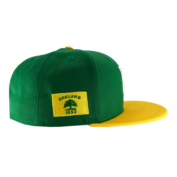A green and yellow New Era Oakland Ballers 59Fifty Cap by Oakland Ballers is depicted. The back panel is green with a yellow rectangular patch that displays a green tree and the text "OAKLAND 1852," capturing the essence of Oakland sports team pride. The brim of the cap is yellow. The hat is presented on a white background, highlighting its side and back views.