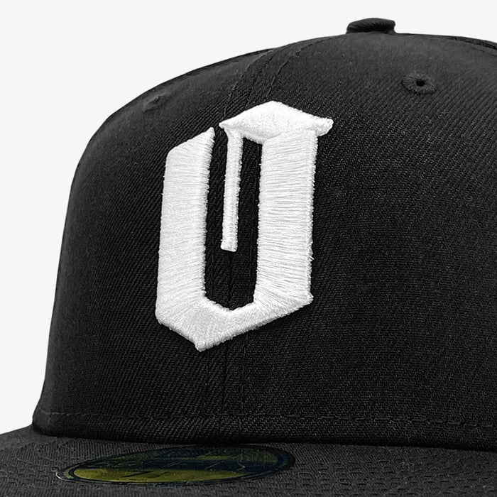 Close-up of a black New Era Official 59FIFTY Fitted Cap by Other, showcasing a large, bold, white embroidered “U” logo on the front panel. A textured authenticity sticker is visible on the brim. The background is plain white.