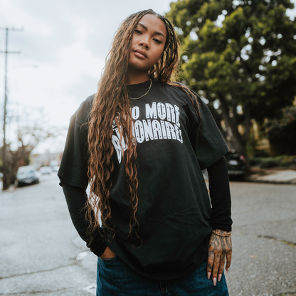 A young person with long braids confidently stands on a quiet street wearing the BOSK No Billionaires Tee, a classic heavy-weight cotton shirt. Under an overcast sky with trees and parked cars in the background, tattoos adorn their hand.