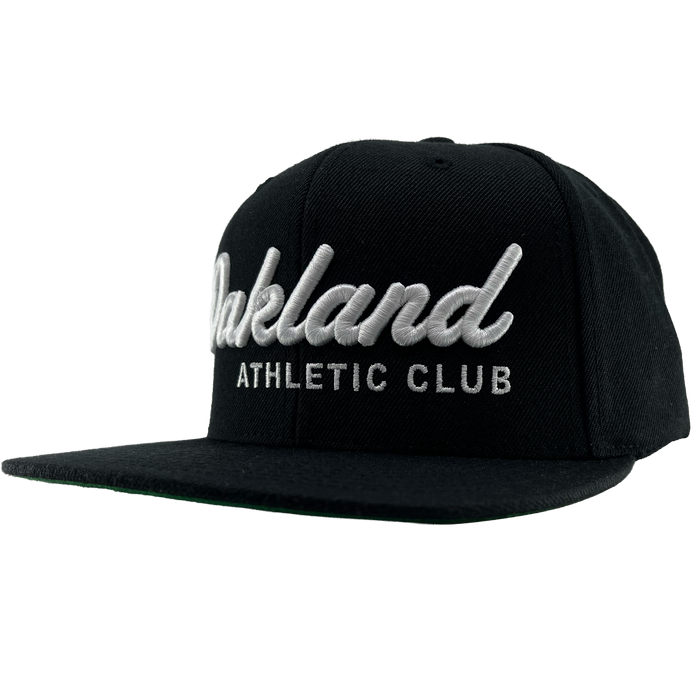 The OAC Script Snapback by OAC is a black snapback cap featuring white embroidered “Oakland” in cursive with “ATHLETIC CLUB” in bold capital letters beneath it. This adjustable hat is displayed at a slight angle on a plain white background, highlighting the classic style of the Oakland Athletic Club.