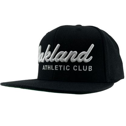 The OAC Script Snapback by OAC is a black snapback cap featuring white embroidered “Oakland” in cursive with “ATHLETIC CLUB” in bold capital letters beneath it. This adjustable hat is displayed at a slight angle on a plain white background, highlighting the classic style of the Oakland Athletic Club.