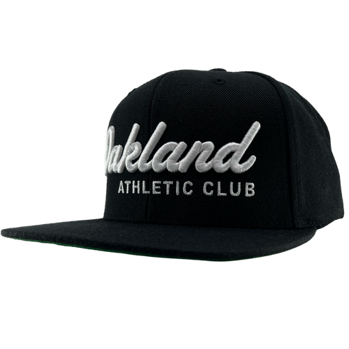 The OAC Script Snapback by OAC is a black snapback cap featuring white embroidered “Oakland” in cursive with “ATHLETIC CLUB” in bold capital letters beneath it. This adjustable hat is displayed at a slight angle on a plain white background, highlighting the classic style of the Oakland Athletic Club.