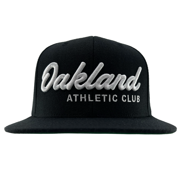 The OAC Script Snapback by OAC is a black flat-brimmed, adjustable cap featuring bold white cursive “Oakland” lettering above smaller block “ATHLETIC CLUB” text on the front, standing out against a plain white background.