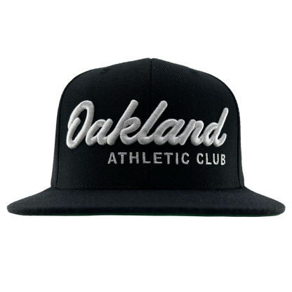 The OAC Script Snapback by OAC is a black flat-brimmed, adjustable cap featuring bold white cursive “Oakland” lettering above smaller block “ATHLETIC CLUB” text on the front, standing out against a plain white background.