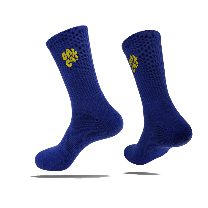 Side view of a pair of blue crew socks with a yellow embroidered OAK CA blob-style wordmark on the side calves.