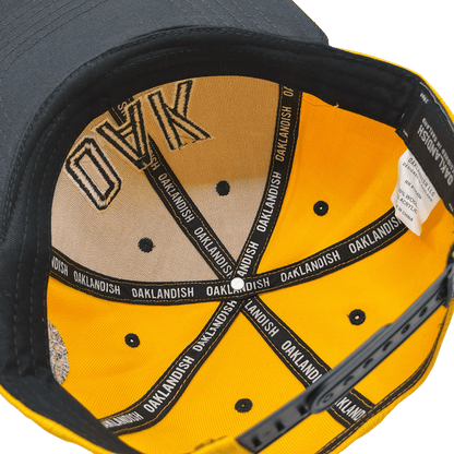 The image shows the inside of an OAK Block Snapback by Oaklandish, which has a black exterior and yellow lining. The headwear features "OAK" written on the inner part of the brown headband, and its seams repeatedly display "OAKLANDISH" in white text on black fabric strips. Eyelets are visible around the inner crown.