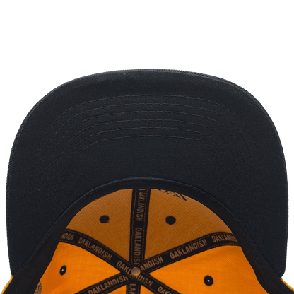 A close-up view of the underside of an OAK Block Snapback by Oaklandish shows a black brim with an orange interior featuring black trim. The word "OAKLANDISH" is repeatedly printed in yellow along the black seams.