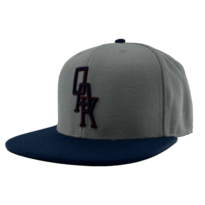 The OAK Monogram Snapback by Oaklandish features a gray crown with a flat, navy blue brim. An embroidered black OAK logo with a red outline adorns the front, and the top button matches in navy blue. Displayed from a front-angled perspective, this hat stylishly celebrates Oaklandish pride.