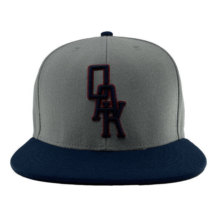 A close-up image of the OAK Monogram Snapback by Oaklandish, showcasing its grey body and navy blue brim. Embroidered in navy blue and outlined in red on the front panel is the distinctive "OAK" logo, evoking Oaklandish style. The cap is centered against a plain green background.