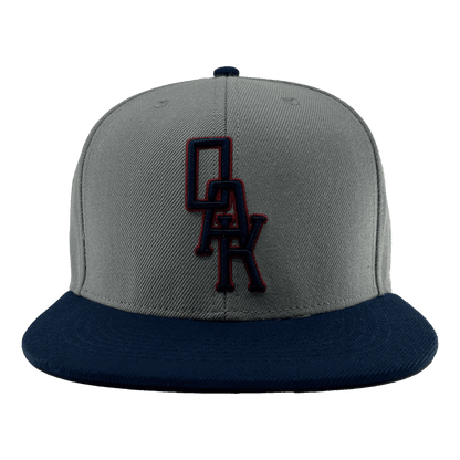 A close-up image of the OAK Monogram Snapback by Oaklandish, showcasing its grey body and navy blue brim. Embroidered in navy blue and outlined in red on the front panel is the distinctive "OAK" logo, evoking Oaklandish style. The cap is centered against a plain green background.