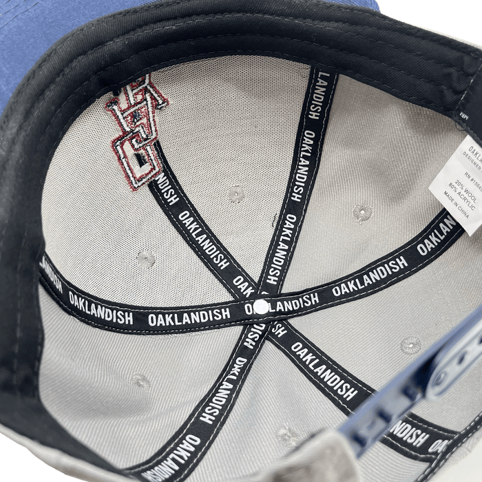 The inside of the OAK Monogram Snapback is shown, featuring light gray fabric with black reinforcing tapes. The tapes read "OAKLANDISH" repeatedly in white text. There is a label with washing instructions and size information attached. Additionally, part of an embroidered OAK logo is visible on the fabric.