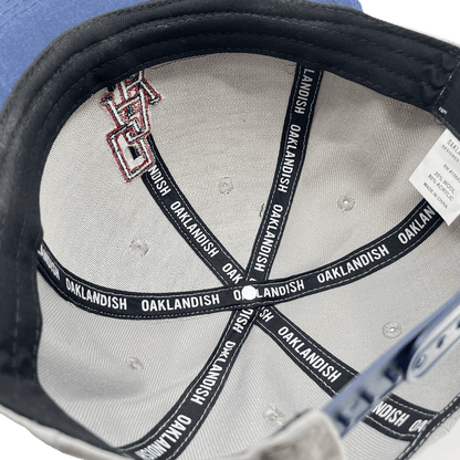 The inside of the OAK Monogram Snapback is shown, featuring light gray fabric with black reinforcing tapes. The tapes read "OAKLANDISH" repeatedly in white text. There is a label with washing instructions and size information attached. Additionally, part of an embroidered OAK logo is visible on the fabric.
