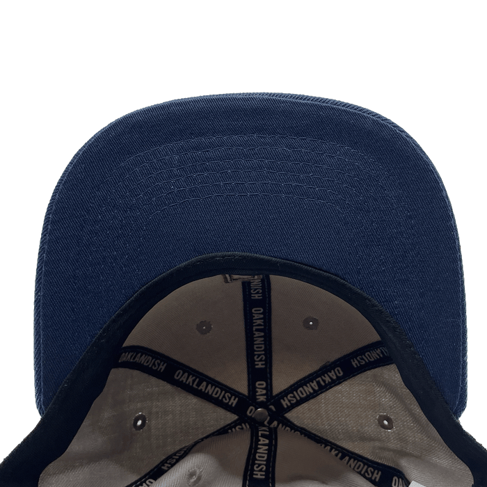The OAK Monogram Snapback from Oaklandish is viewed from above, showcasing the brim and interior. The inside features white fabric with black edges and adjustable straps. The straps are printed with the word "Oaklandish" in white text on a blue background. This clean snapback hat also boasts an embroidered OAK logo on the front.
