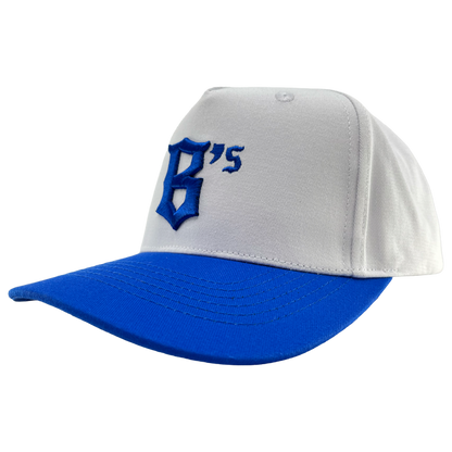 The Oakland Ballers 5 Panel Cap is a white snapback hat with a royal blue brim. The front showcases a bold, stylized blue “B” alongside a small blue “‘s,” representing the Oakland Ballers. The background of the cap is plain white.