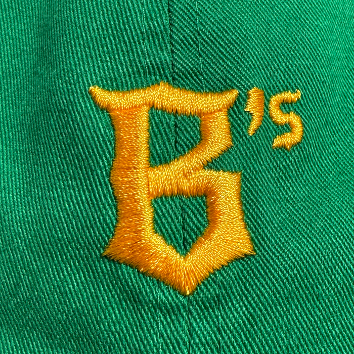 Close-up of the Oakland Ballers Dad Hat by Oakland Ballers, featuring green fabric with yellow embroidery: a large, stylized "B" and a smaller superscript "’s". Detailed stitching highlights the visible texture of the hat.
