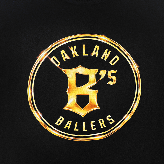 The Oakland Ballers Gold Tee features a circular gold emblem on black with "OAKLAND BALLERS" encircling a bold “B” and smaller "'S", highlighted by radiant metallic gold reflections. Brand: Oakland Ballers.