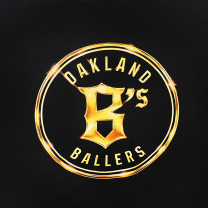 The Oakland Ballers Gold Tee features a circular gold emblem on black with "OAKLAND BALLERS" encircling a bold “B” and smaller "'S", highlighted by radiant metallic gold reflections. Brand: Oakland Ballers.