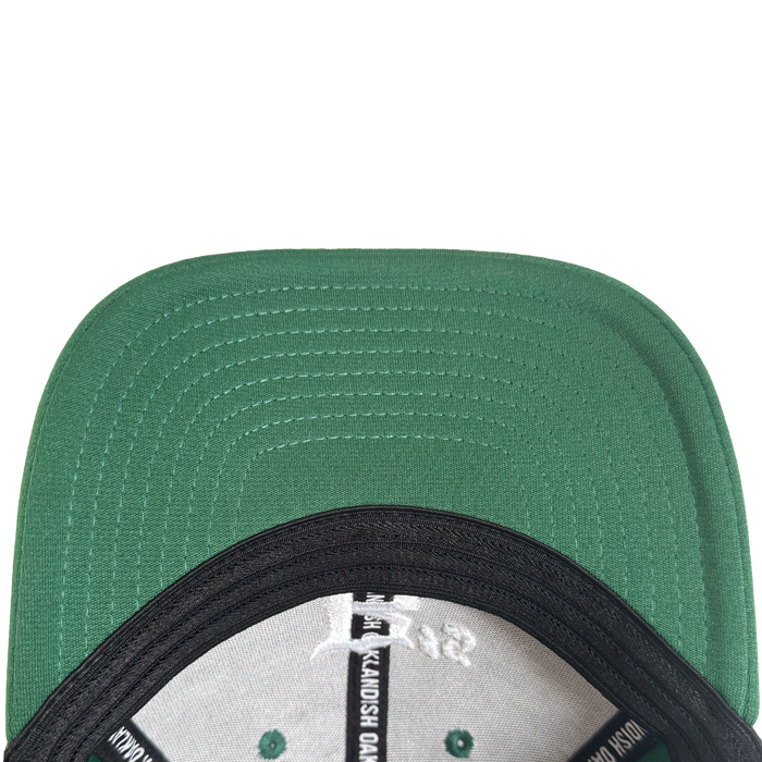 Close-up, top-down view of the Oakland Ballers Logo Flexfit Hat. The green flat brim shows stitched lines underneath, and inside are black and white labels with seams. The background is white. Brand: Oakland Ballers.