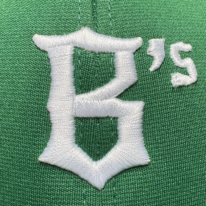 Close-up of the Oakland Ballers Logo Flexfit Hat by Oakland Ballers, featuring green fabric with a large, stylized white embroidered "B," plus a raised white apostrophe and "s," highlighting the hat’s detailed stitching and rich texture.