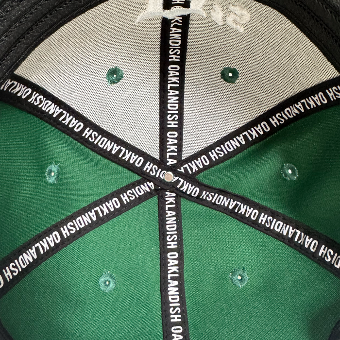 View of the inside top of an Oakland Ballers Logo Flexfit Hat by Oakland Ballers. Black and white taping says "OAKLANDISH" along the seams, with green stitched ventilation holes, celebrating local baseball team pride.