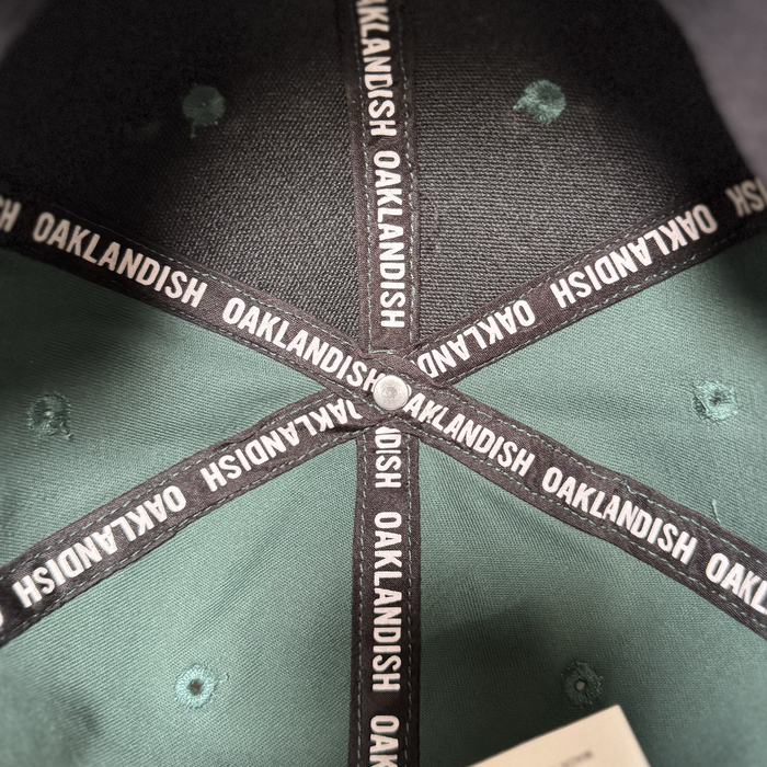 Close-up of the Oakland Ballers Logo Hat’s interior reveals black fabric strips with "OAKLANDISH" in white, celebrating the Oakland Ballers baseball team. The green and black panels meet at the top, secured by a metal rivet.