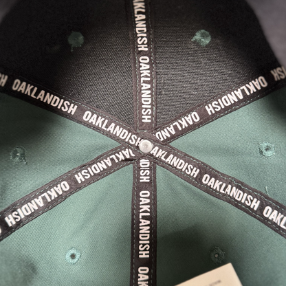 Close-up of the Oakland Ballers Logo Hat’s interior reveals black fabric strips with "OAKLANDISH" in white, celebrating the Oakland Ballers baseball team. The green and black panels meet at the top, secured by a metal rivet.