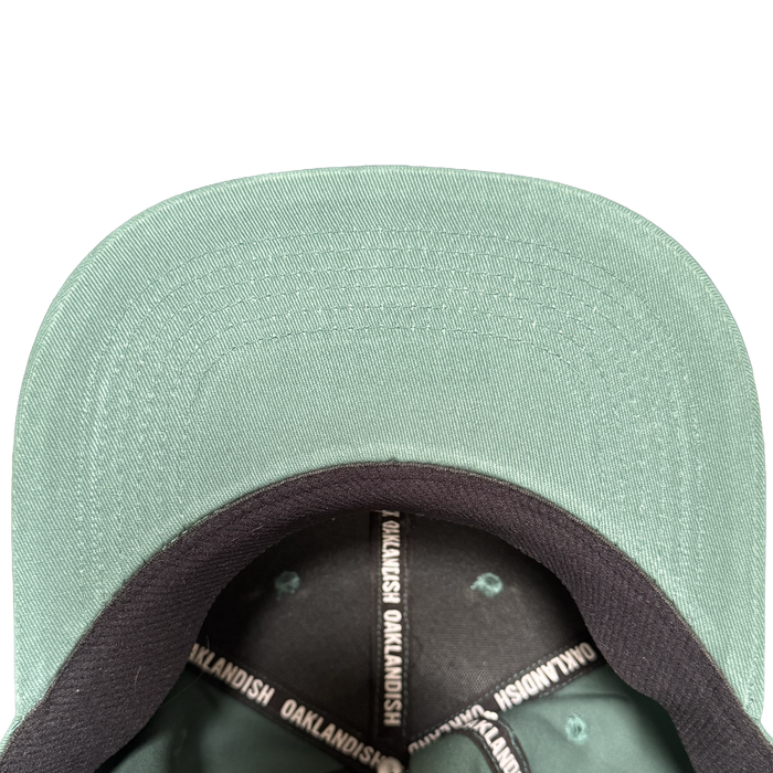 Close-up of the Oakland Ballers Logo Hat by Oakland Ballers, shown from above with focus on the green brim’s underside and black inner band printed with “OAKLANDISH” in white—a must-have for any Oakland baseball fan. Plain white background.
