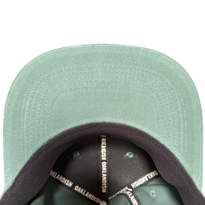 Close-up of the Oakland Ballers Logo Hat by Oakland Ballers, shown from above with focus on the green brim’s underside and black inner band printed with “OAKLANDISH” in white—a must-have for any Oakland baseball fan. Plain white background.