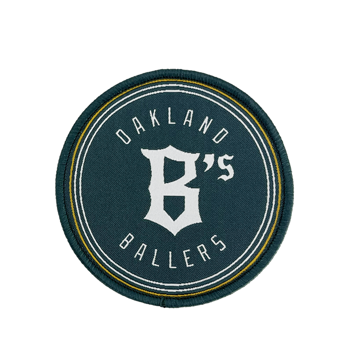 The Oakland Ballers Logo Patch from the Oakland Ballers features a circular design with a dark green background. The words "Oakland Ballers" are arched along the top and bottom edges, while a large, stylized white "B's" occupies the center. Representing the professional baseball team in the Pioneer League, this patch is bordered by thin white and yellow rings.
