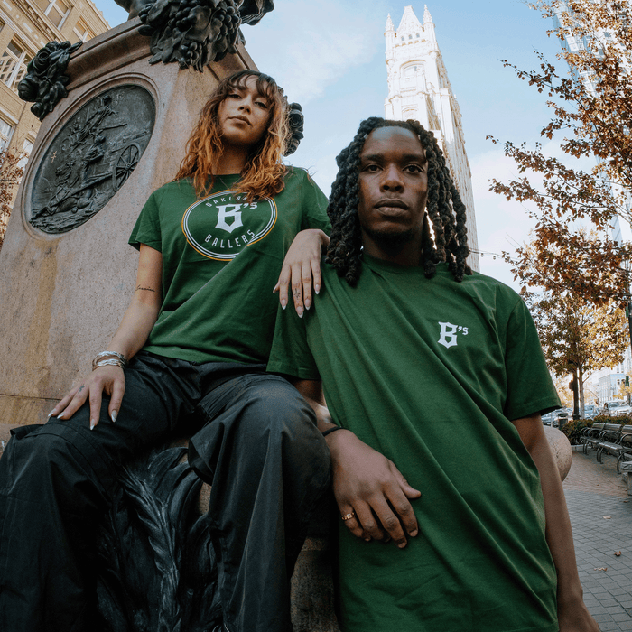Two individuals pose in front of a city monument. The person on the left is seated with a hand on the monument, wearing an Oakland Ballers Logo Tee and black pants, and has curly hair. The person on the right stands, also in a green T-shirt with "B's" logo and long braided hair. A tall building and autumn trees create a picturesque backdrop evocative.