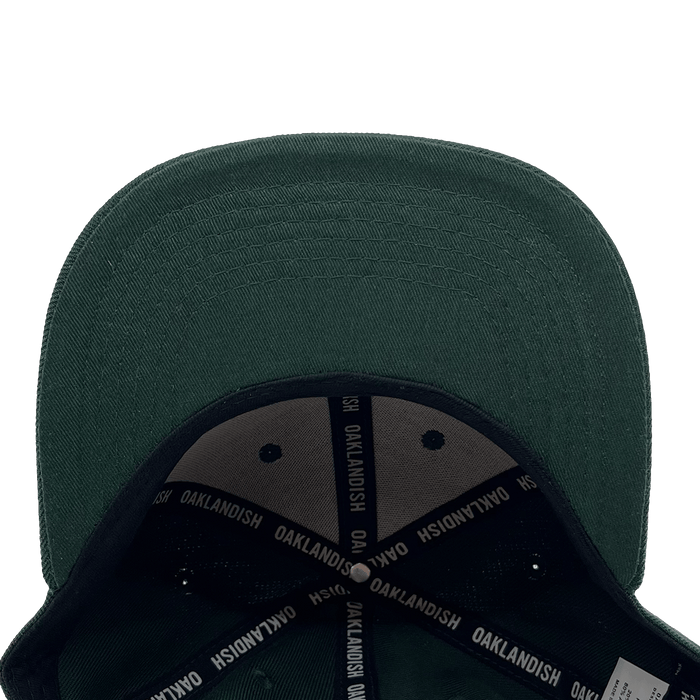 An Oakland Ballers Snapback in green, seen from above, displaying its wide, slightly curved brim. The underside showcases black fabric with white "OAKLANDISH" text repeatedly printed on the inner seams. The cap's interior structure reveals a partial view of a label referencing the Oakland Ballers professional baseball team.