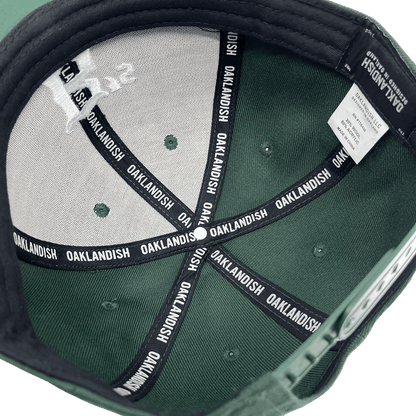 Interior view of the Oakland Ballers Snapback. The inside features black seams repeatedly labeled "OAKLANDISH" in white text. The label inside includes washing instructions and material information. The cap also has a small logo design on the inner top panel, celebrating the Oakland Ballers, a Pioneer League professional baseball team. The brim is lined with black fabric.