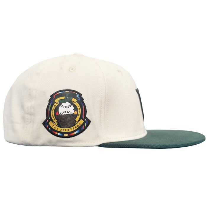 A white Oakland Peloteros Hat by the Oakland Ballers is displayed from a side view. The hat features a green brim and a colorful embroidered patch with a stylized baseball, crossed bats, and intricate detailing—a tribute to the team as an exciting addition to the Pioneer League. The plain white background emphasizes the vibrant colors and design of the cap.