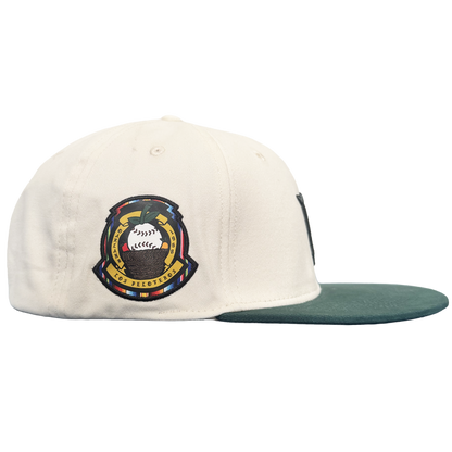 A white Oakland Peloteros Hat by the Oakland Ballers is displayed from a side view. The hat features a green brim and a colorful embroidered patch with a stylized baseball, crossed bats, and intricate detailing—a tribute to the team as an exciting addition to the Pioneer League. The plain white background emphasizes the vibrant colors and design of the cap.