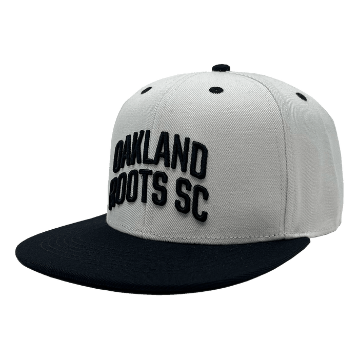 The Oakland Roots SC Arched Text Snapback by Oakland Roots SC features a white crown complemented by a black flat brim and matching black ventilation eyelets. The front is adorned with raised embroidery of the text "OAKLAND ROOTS SC," exuding a sleek, minimalistic design that perfectly captures the sporty essence of the Oaklandish style.