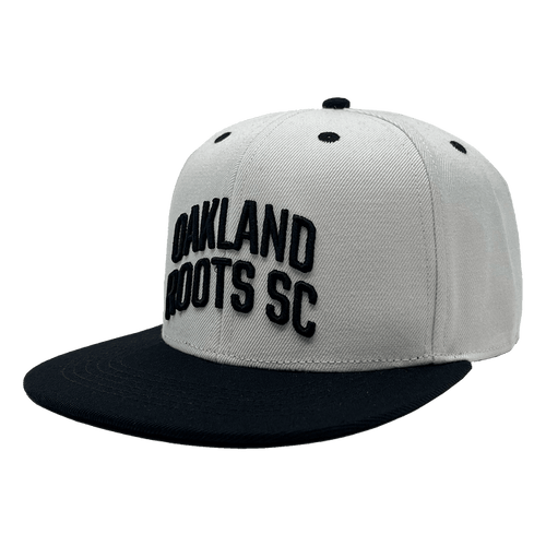 The Oakland Roots SC Arched Text Snapback by Oakland Roots SC features a white crown complemented by a black flat brim and matching black ventilation eyelets. The front is adorned with raised embroidery of the text "OAKLAND ROOTS SC," exuding a sleek, minimalistic design that perfectly captures the sporty essence of the Oaklandish style.
