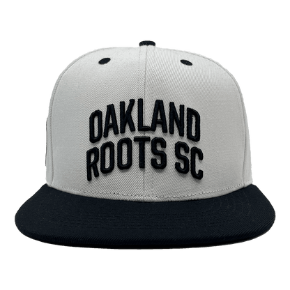 Introducing the Oakland Roots SC Arched Text Snapback by Oakland Roots SC: a gray snapback hat with a black brim. The front prominently displays "OAKLAND ROOTS SC" in bold, black, raised letters. Two small black eyelets on the top provide ventilation. The overall design is simple and sporty, capturing the essence of Oaklandish spirit that represents the Oakland Roots.